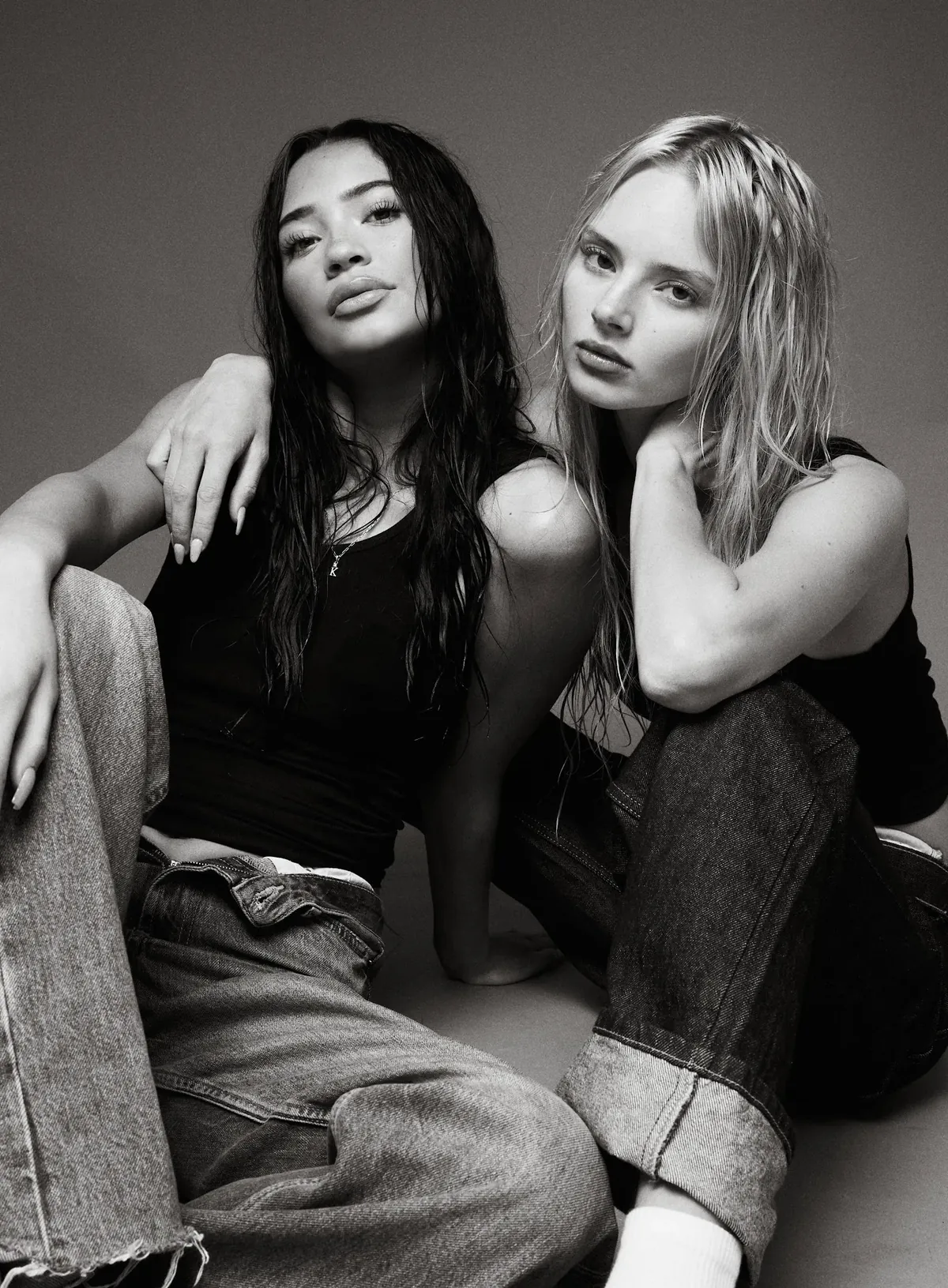 Black-and-white studio portrait of Kaila Triniti seated beside a blonde model, both in black tank tops and loose jeans.