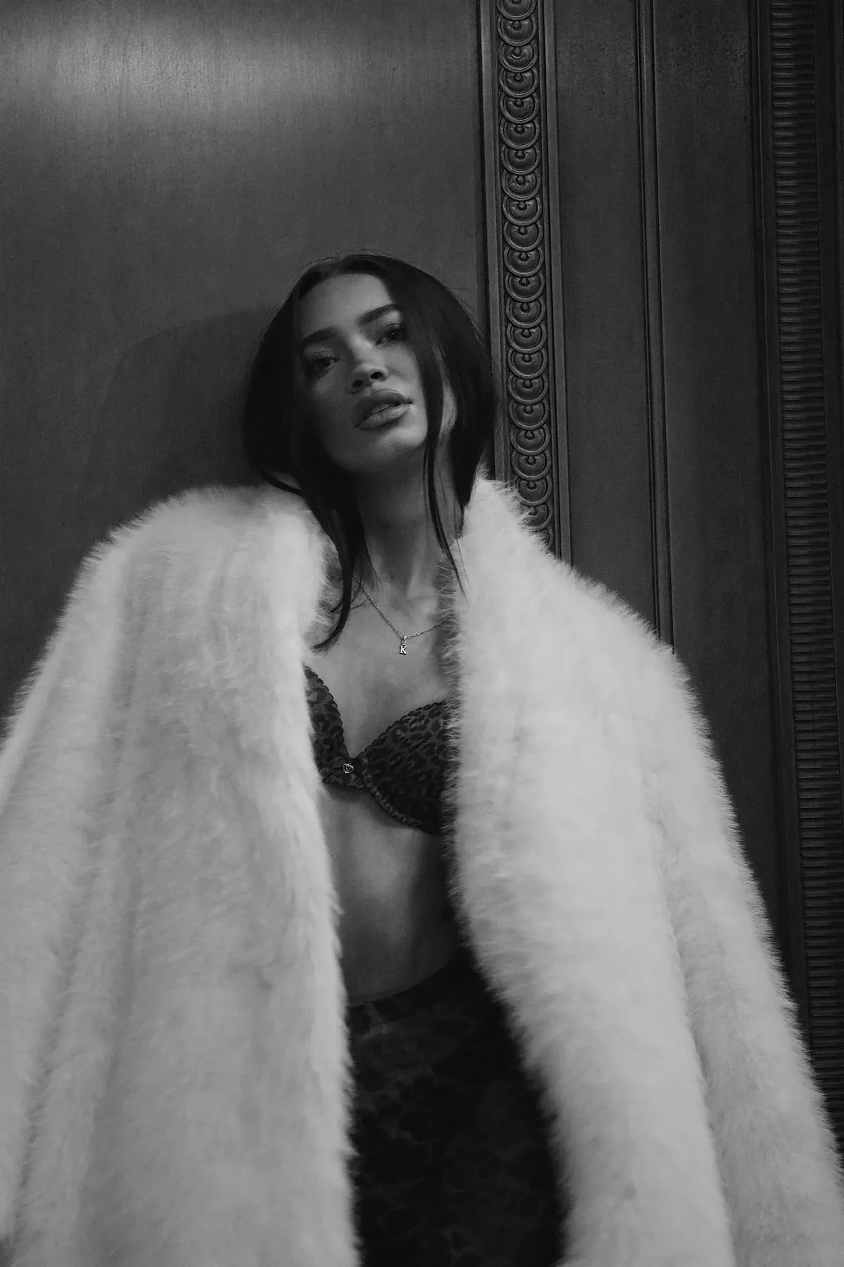 Black-and-white portrait of Kaila Triniti leaning against a carved wooden wall in a white faux-fur coat over a patterned bra.