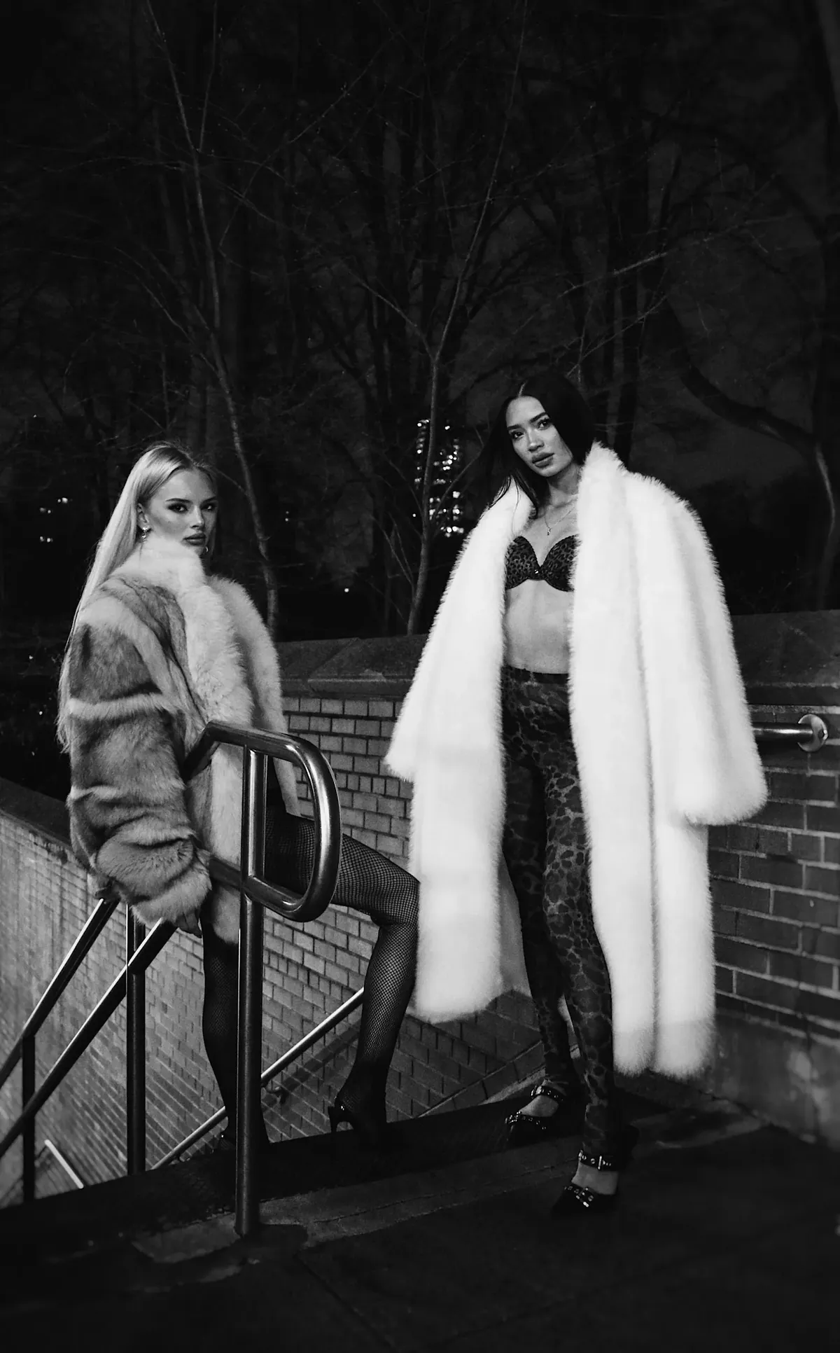 Black-and-white night photograph of Kaila Triniti in a long white faux-fur coat beside another model in fur on an outdoor stairway.