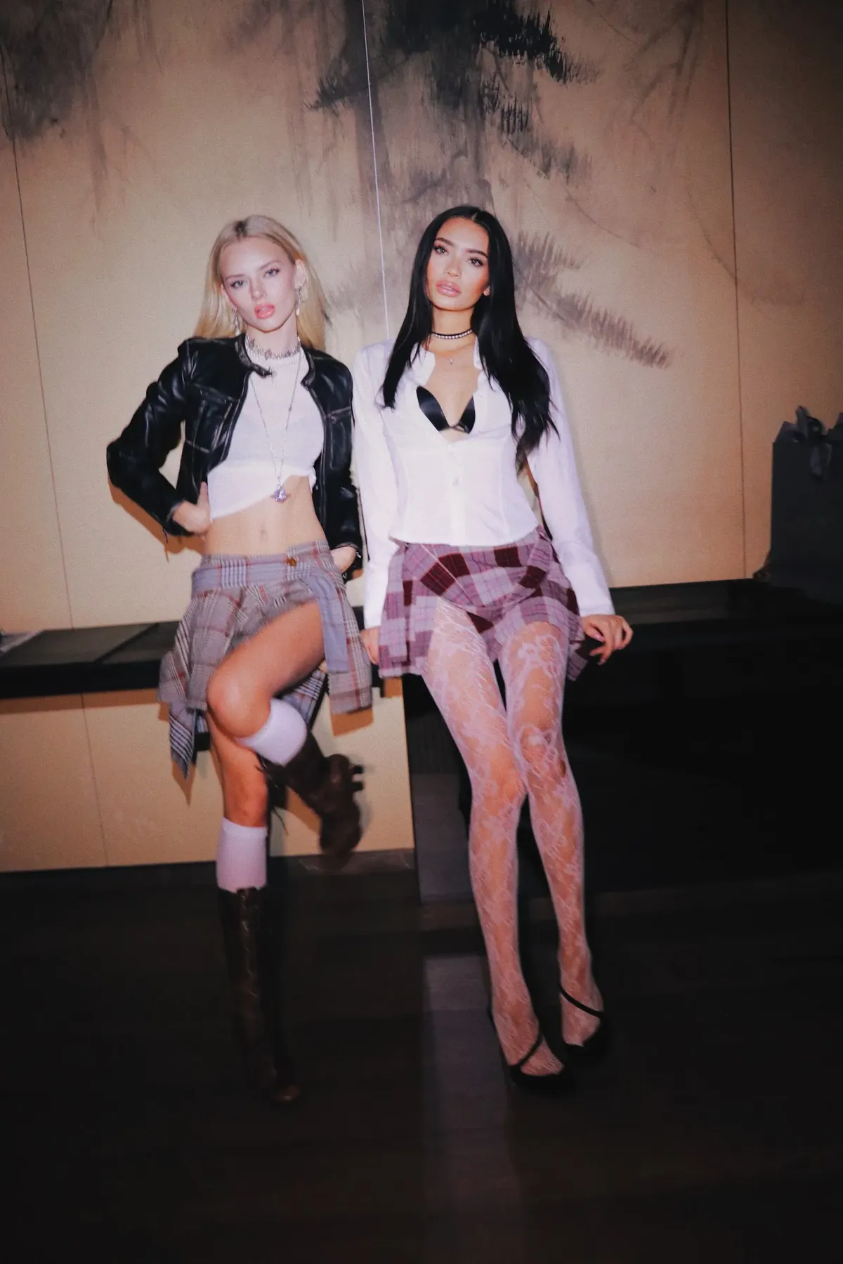 Flash photograph of Kaila Triniti and a blonde model sitting on a ledge in plaid mini skirts and white shirts.