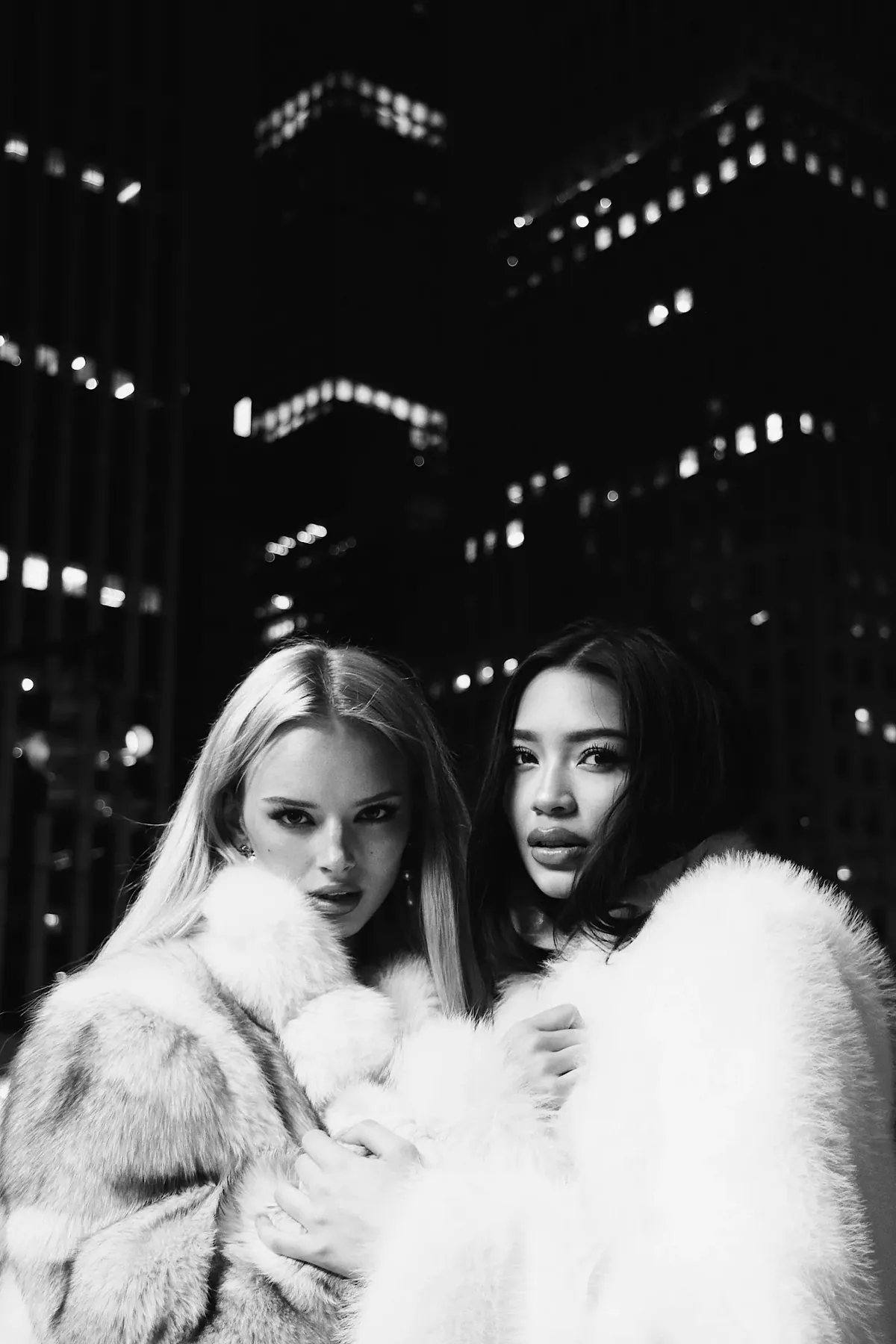 Black-and-white night portrait of Kaila Triniti and a blonde model in fur coats, lit office towers behind them.