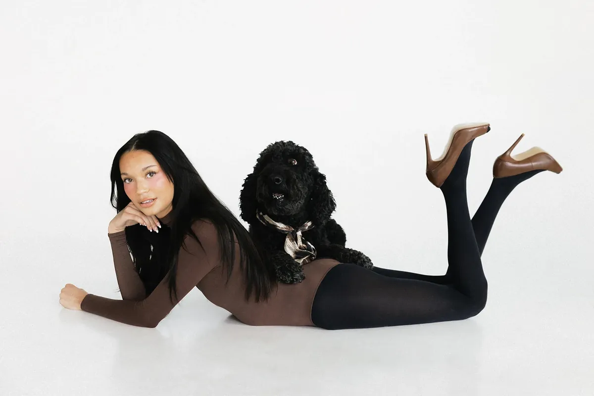 Kaila Triniti lying on her stomach on a white studio floor, chin in hand and cognac heels raised behind her, beside a black curly-coated dog wearing a silk scarf.