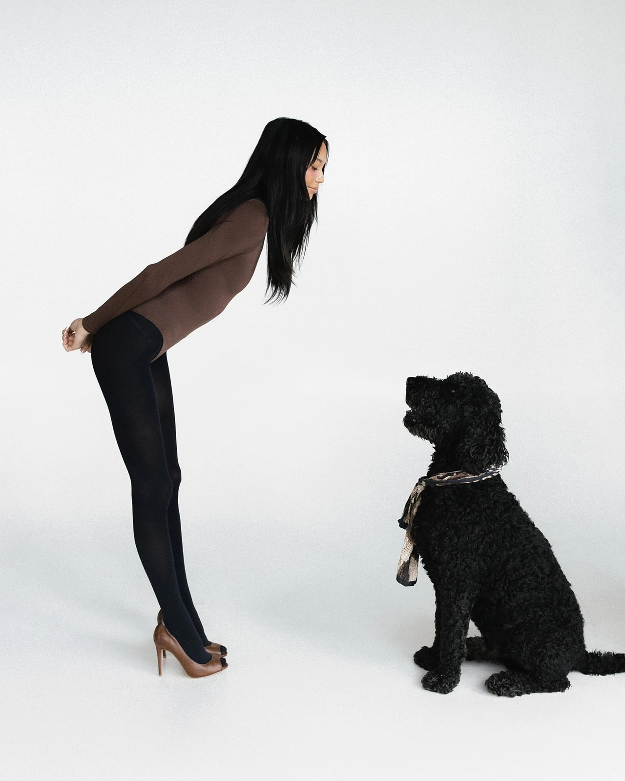 Kaila Triniti in a brown bodysuit, black tights and cognac pumps, bending forward from the hips with her hands clasped behind her back to meet a seated black curly-coated dog wearing a silk scarf.