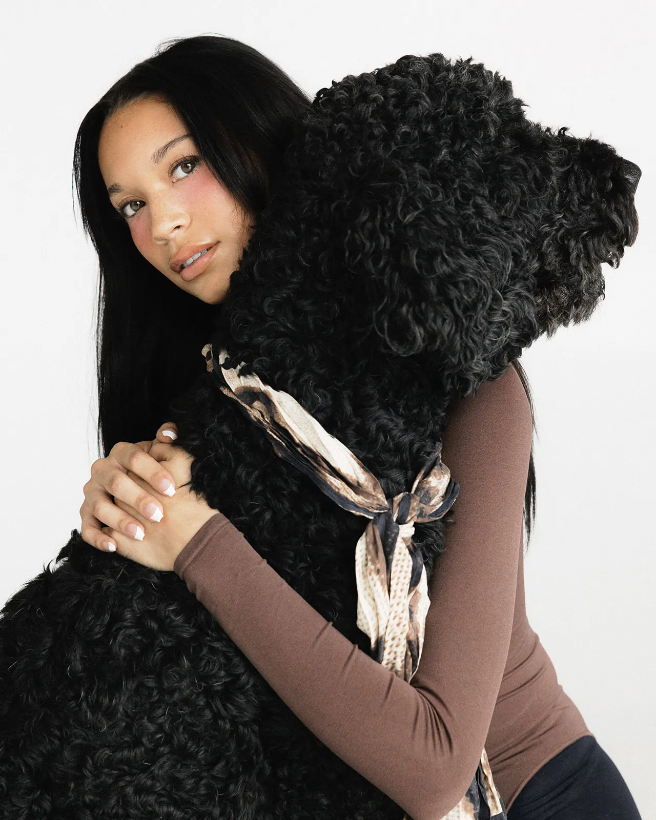 Kaila Triniti hugging a large black curly-coated dog around the neck and looking back over its shoulder at the camera, a printed silk scarf tied at the dog's collar.