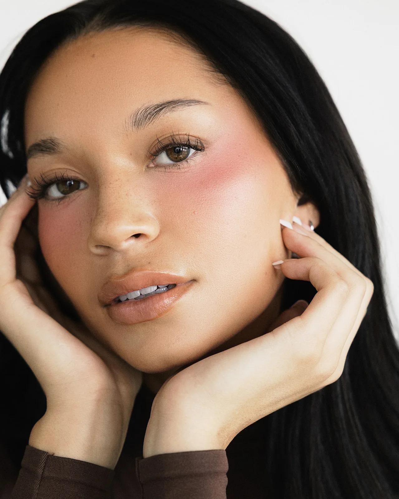 Head-and-shoulders portrait of Kaila Triniti with both hands framing her face, lips slightly parted, long dark hair over her shoulders.