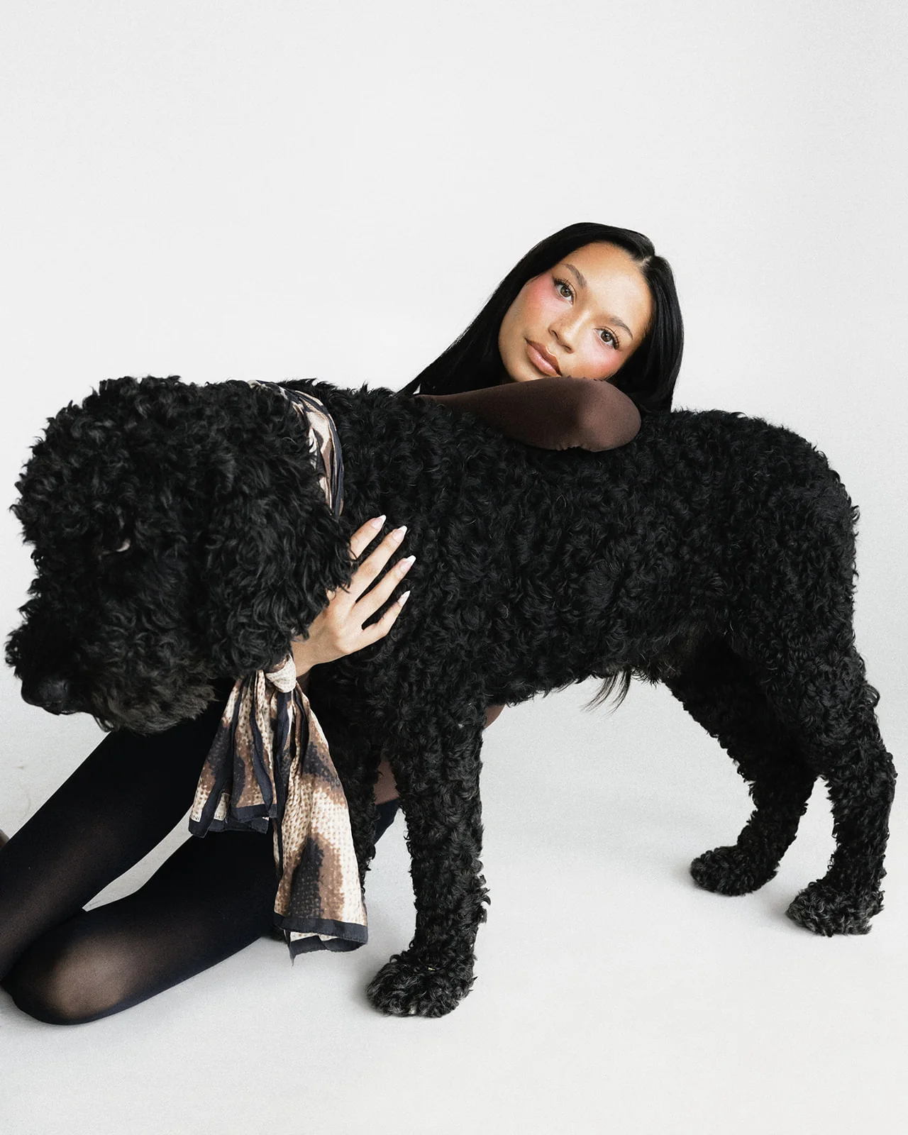 Kaila Triniti kneeling behind a standing black curly-coated dog, one arm folded across its back as a pillow for her head, looking at the camera.