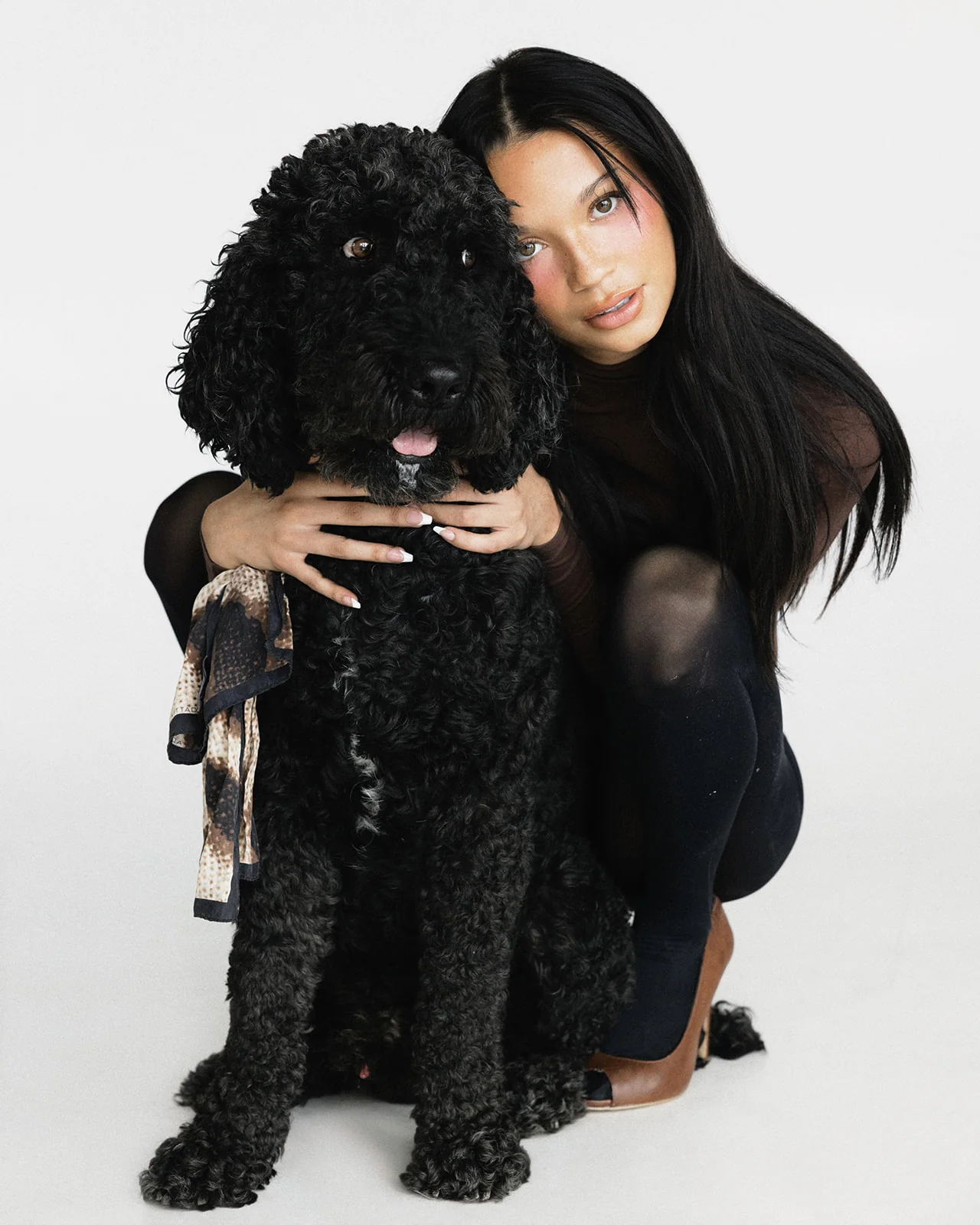 Kaila Triniti crouching behind a seated black curly-coated dog, cheek against its head and hands resting on its chest, with a soft expression toward the camera.