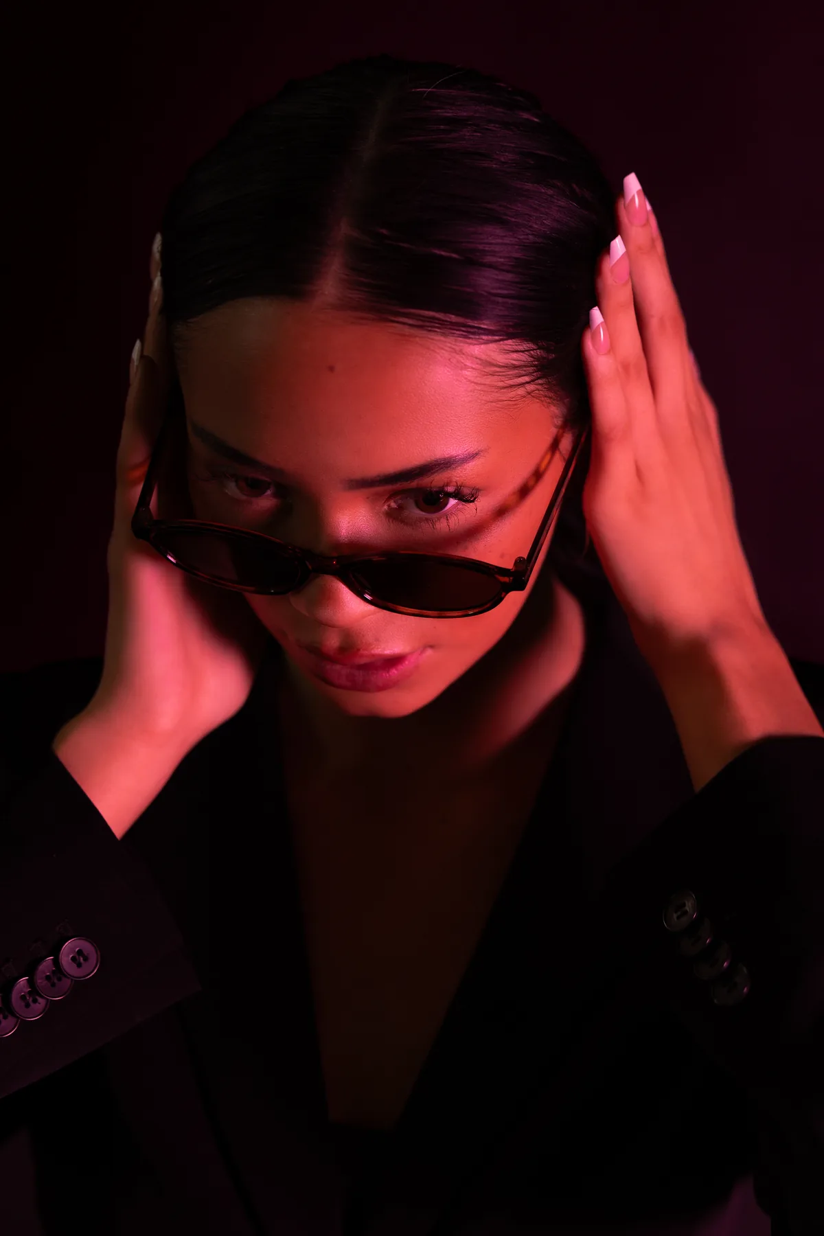 Close portrait of Kaila Triniti under red light, head tilted down, holding her slicked-back hair and looking over tortoiseshell sunglasses.