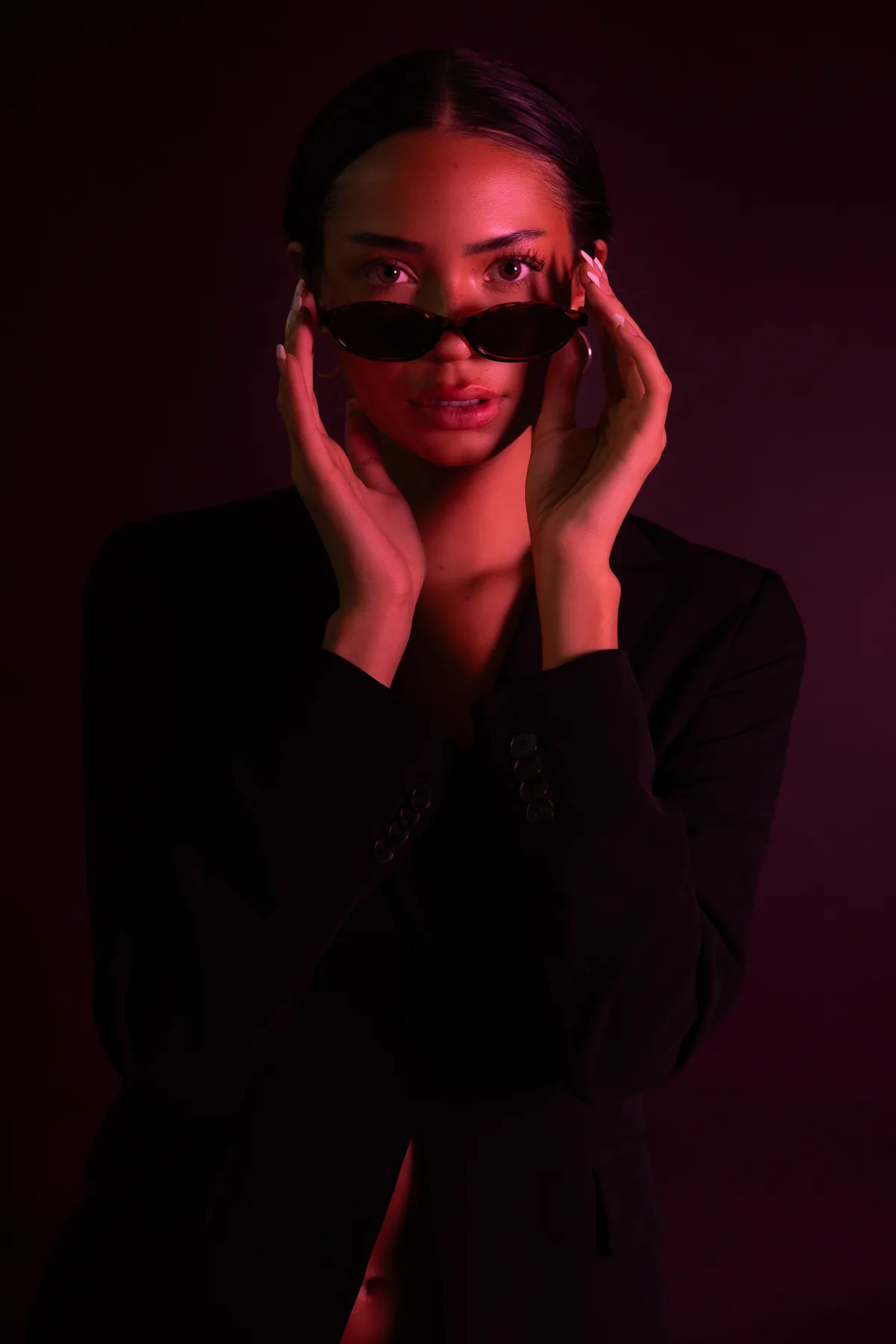 Kaila Triniti in a black blazer under red light, both hands raising oval sunglasses to look straight into the camera.