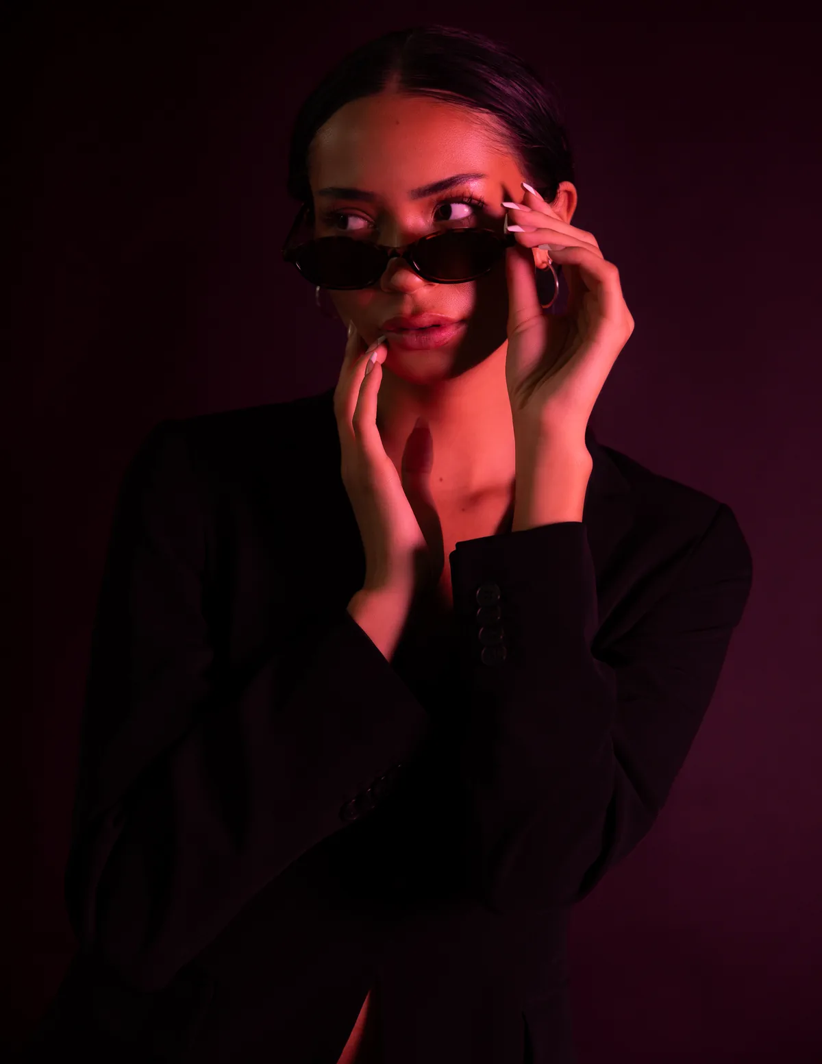 Kaila Triniti in a black blazer under red light, lowering oval sunglasses with one hand and glancing to the side, gold hoop earrings.