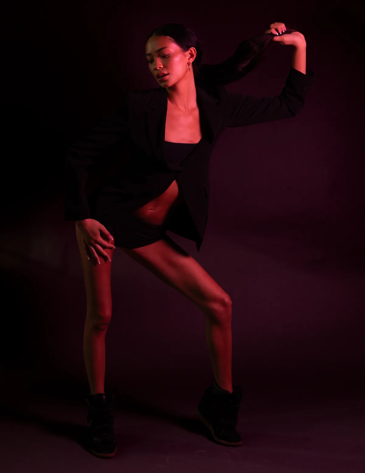 Kaila Triniti in motion under red light, open black blazer and wedge sneakers, one knee bent and one arm pulling her ponytail out to the side.