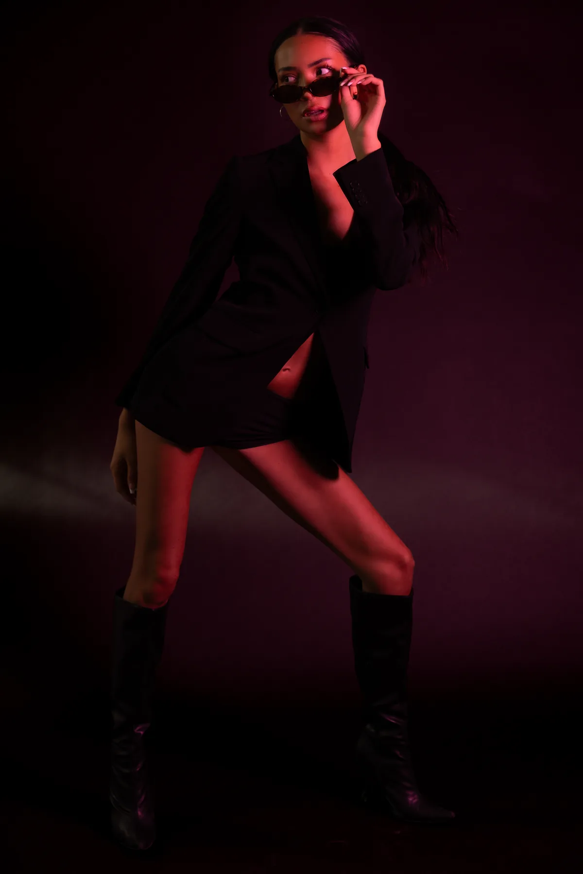 Full-length shot of Kaila Triniti in a black blazer and knee-high boots under red light, stepping wide and glancing back over her sunglasses.