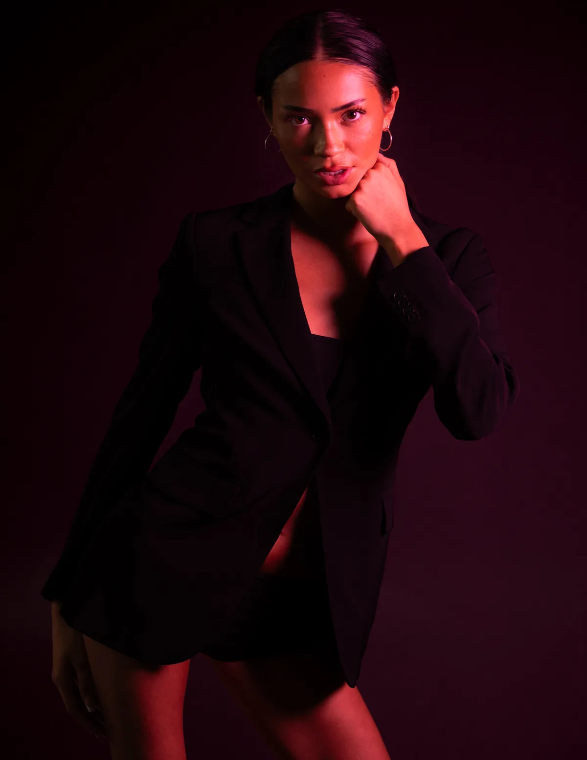 Three-quarter portrait of Kaila Triniti in a black blazer under red light, fist resting at her jaw, looking into the camera.